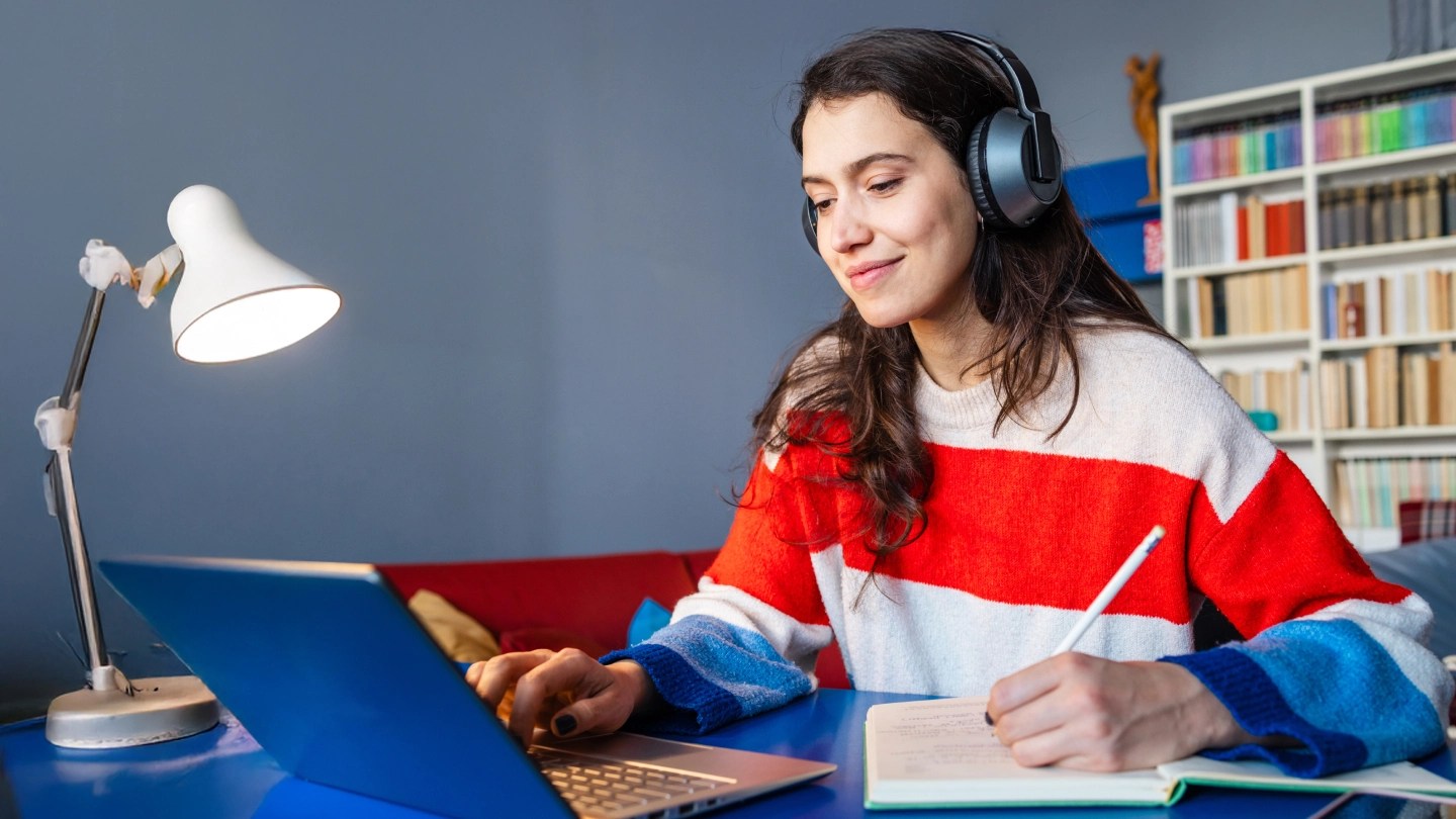 Ragazza con cuffie che prende appunti su un quaderno guardanco un computer