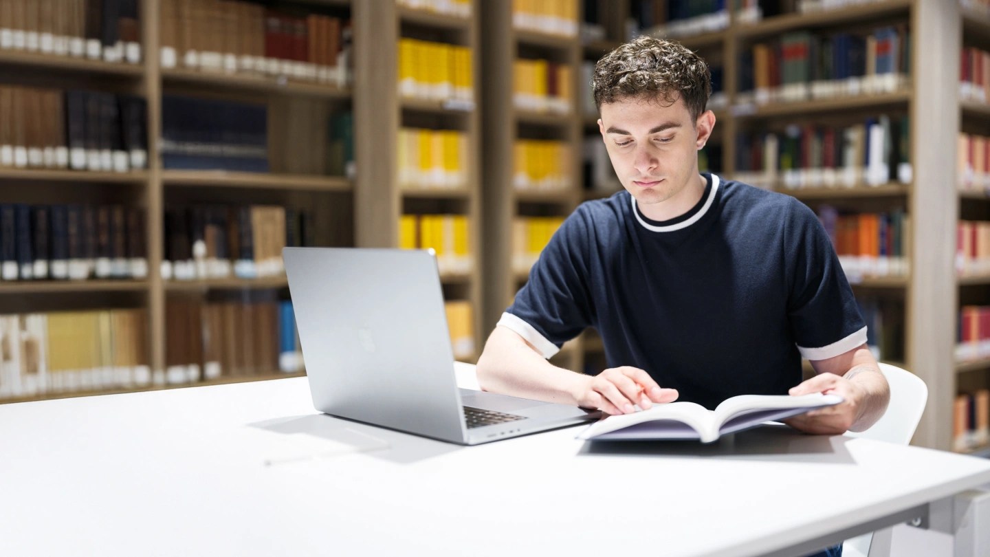 Ragazzo in biblioteca con compueter che sfoglia un libro
