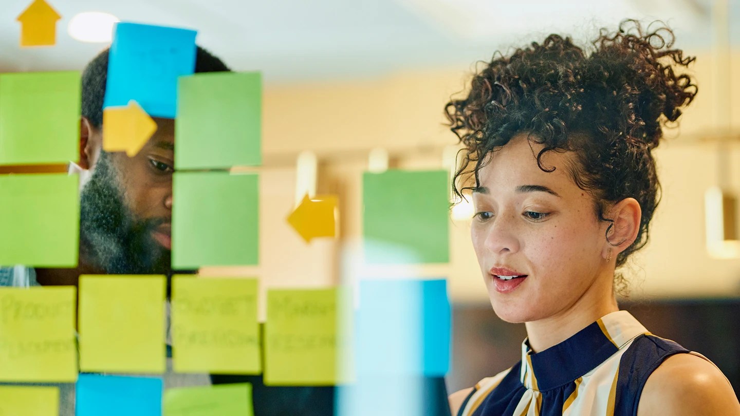 Donna e uomo lavorano al di là di un vetro su cui sono attaccati dei post it colorati